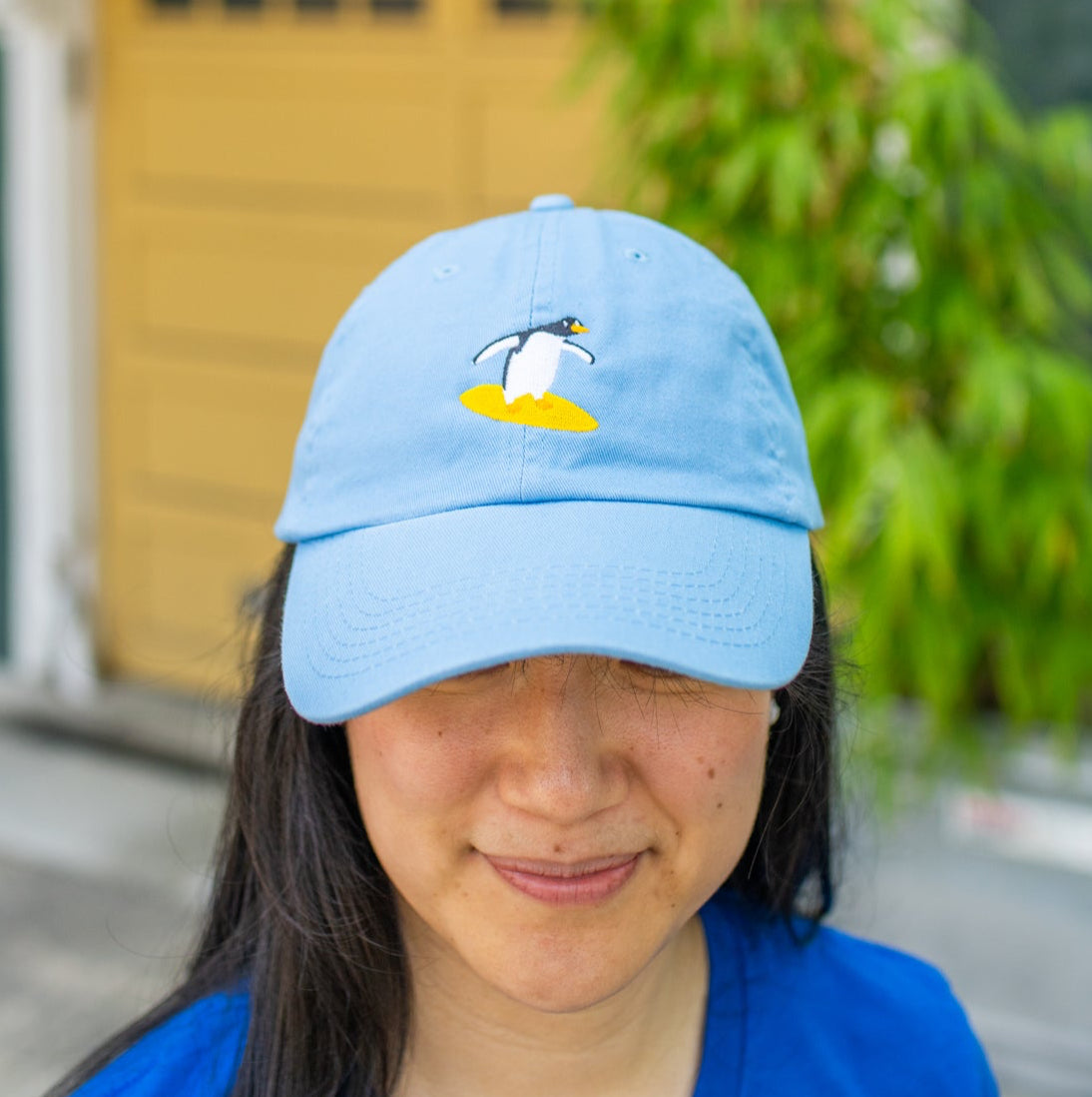 Person wearing sky blue baseball cap with a surfing penguin embroidery and looking down.