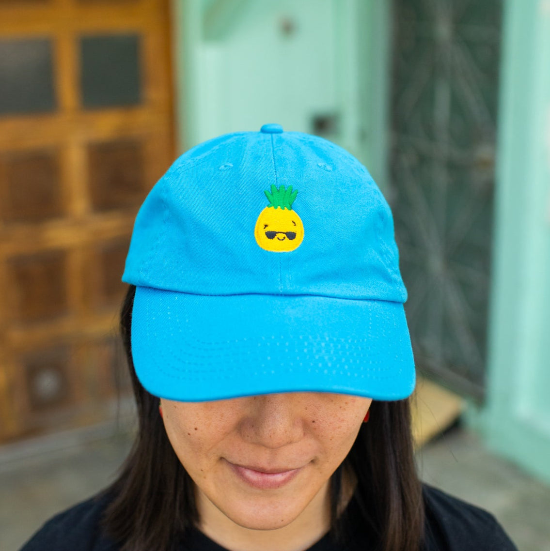 Person wearing bright blue baseball cap with pineapple wearing sunglasses embroidered on the front.