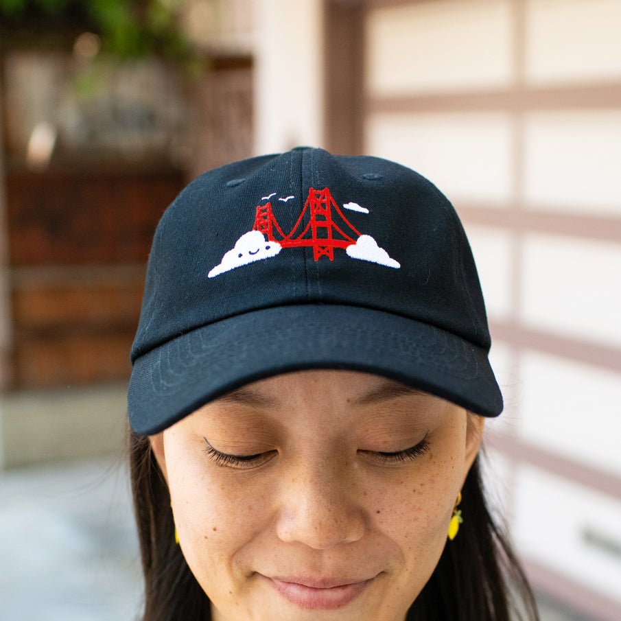 Person smiling wearing black baseball cap with Golden Gate Bridge and smiling clouds embroidery.