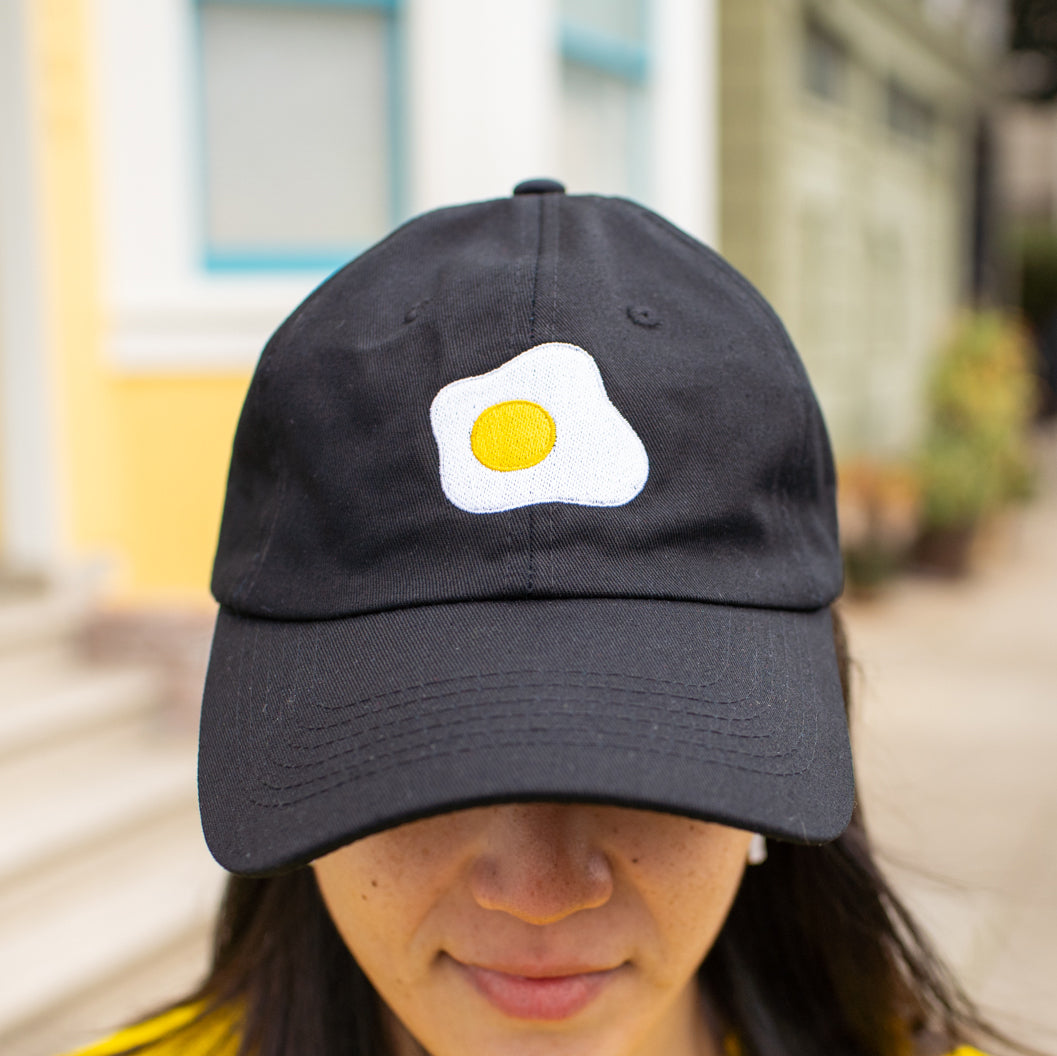 Close-up of person wearing black baseball cap with fried sunny side up egg embroidered on front