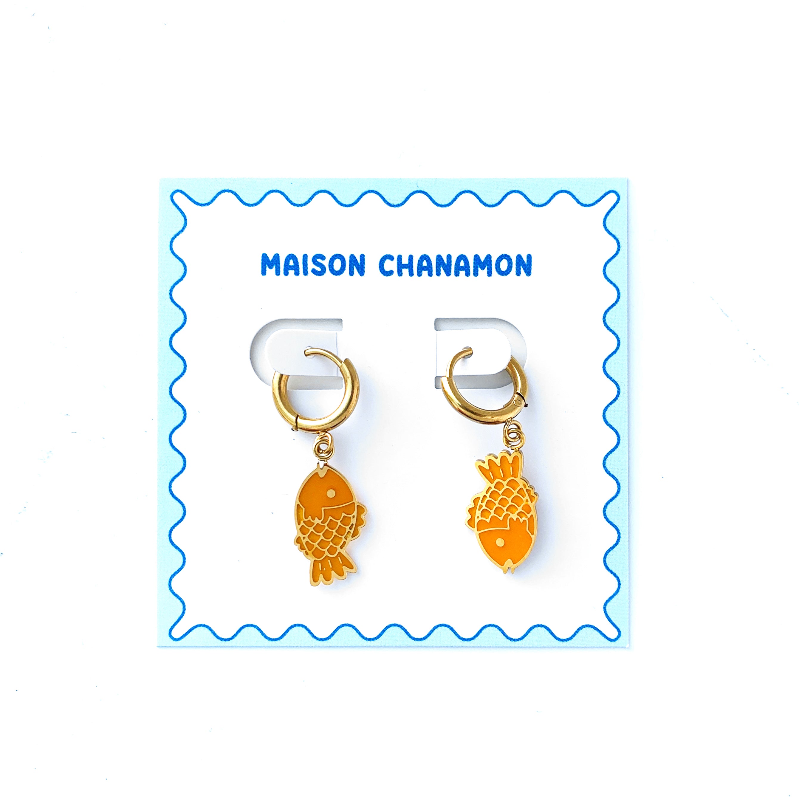 Small gold hoop earrings with charms of a taiyaki fish in opposing directions on a backing card with blue squiggle border and "Maison Chanamon" label.