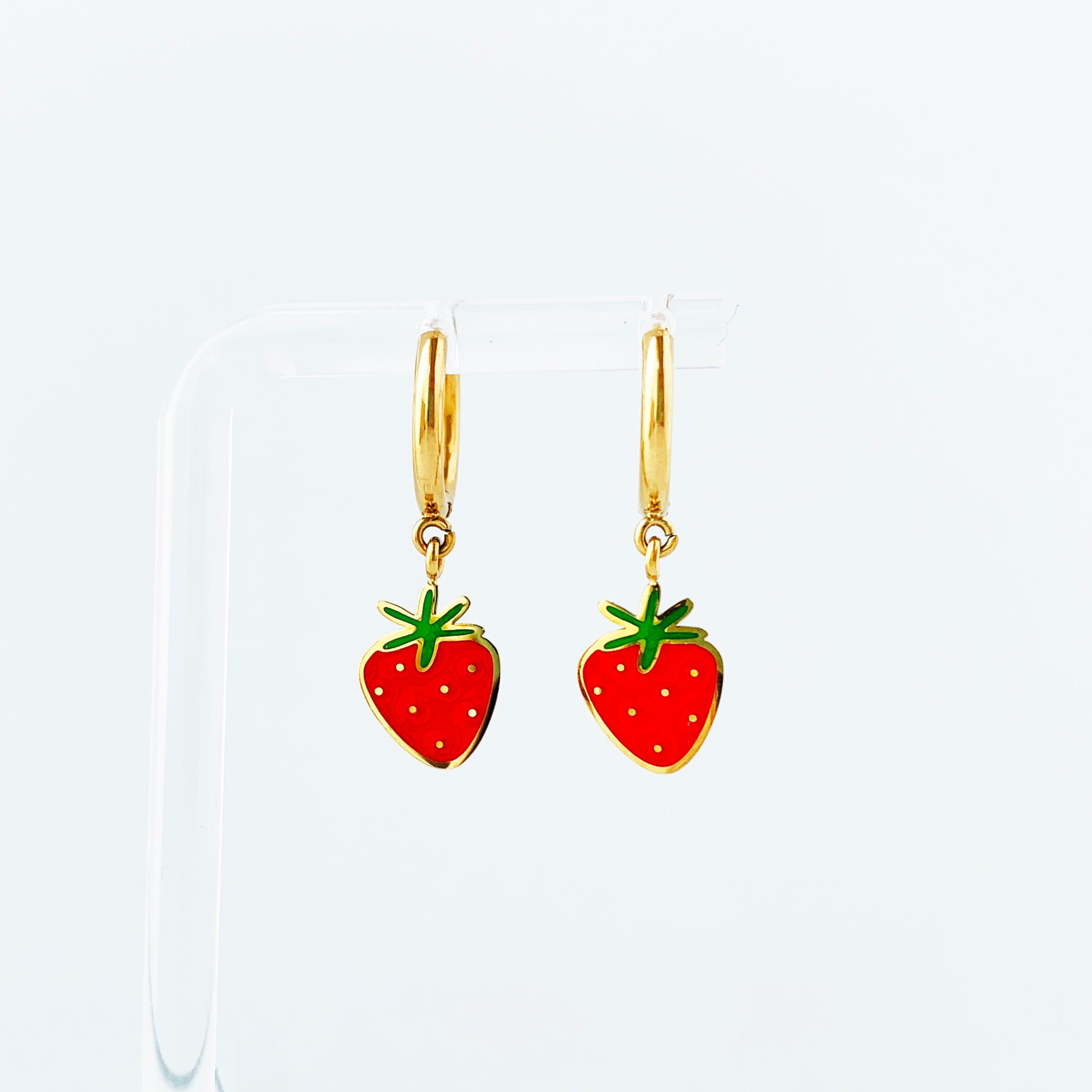 Strawberry enamel charms on gold huggie hoop earrings hanging on a display