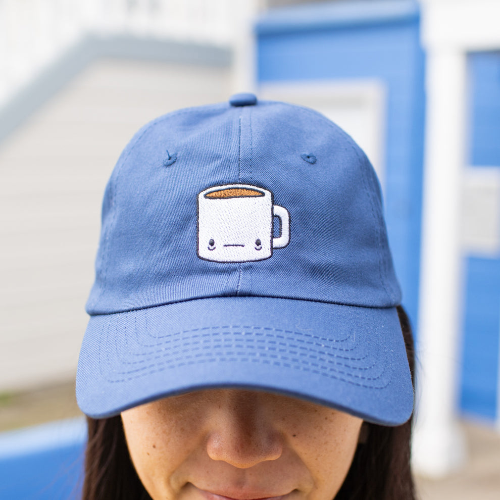 Close up of person wearing navy blue baseball cap with a tired-looking coffee mug embroidered on the front.