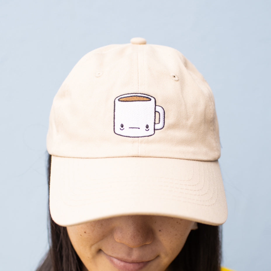 Close up of person wearing beige baseball cap with a tired-looking coffee mug embroidered on the front.