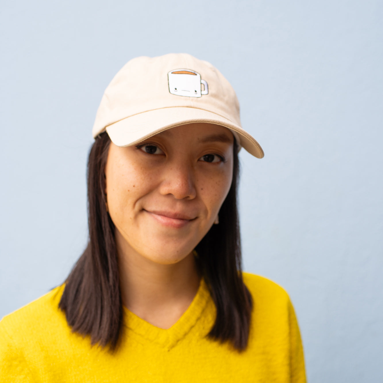 Person wearing beige baseball cap with a tired-looking coffee mug embroidered on the front.