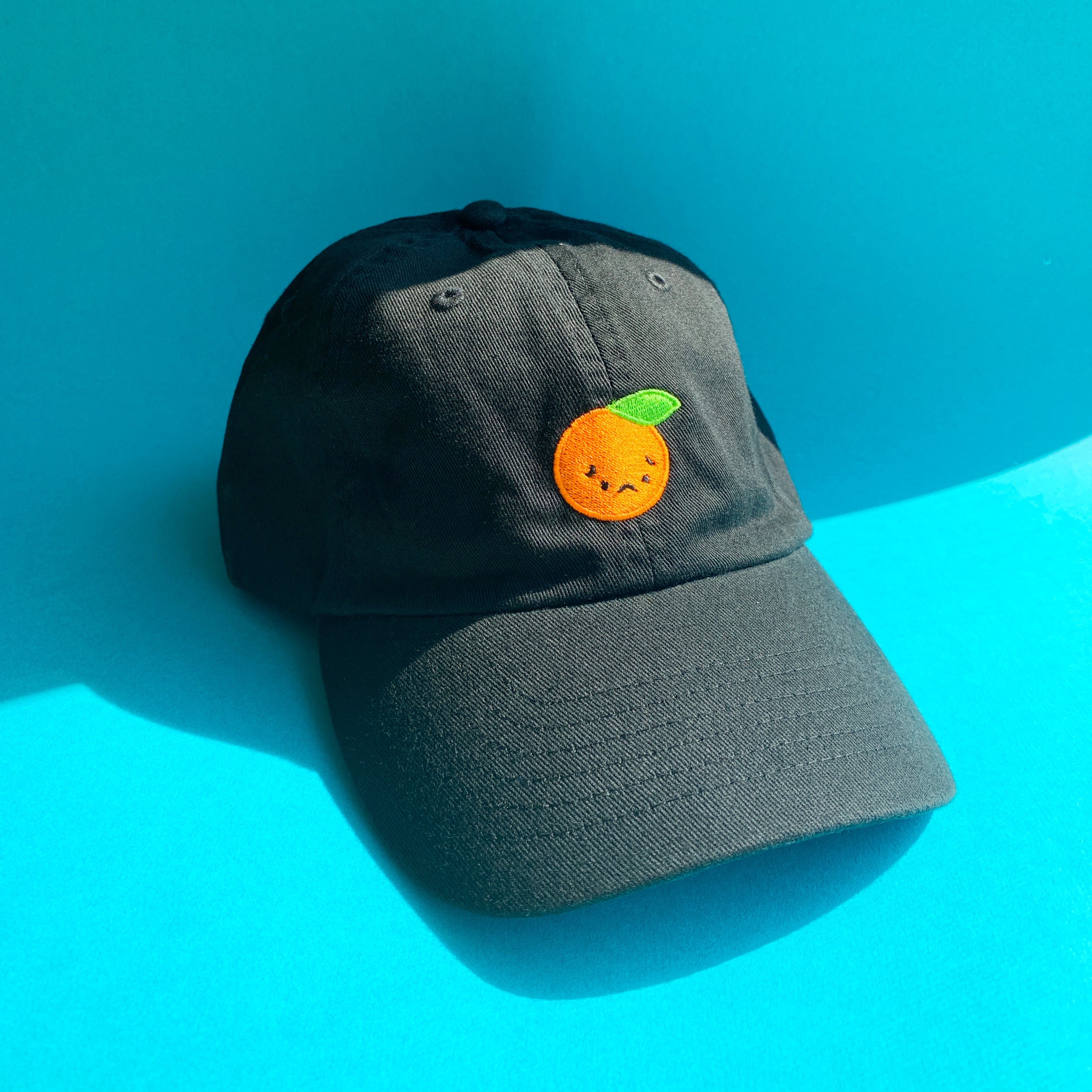 Close up of black baseball cap with sad orange embroidered on front.