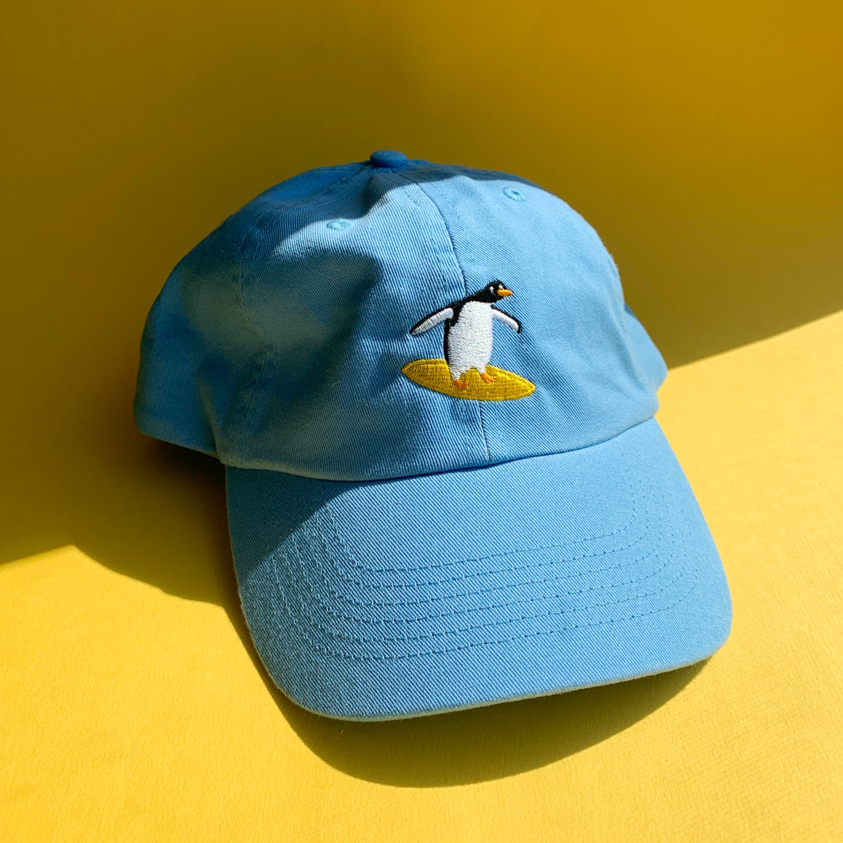 Close up of sky blue baseball cap with a surfing penguin embroidered on the front.
