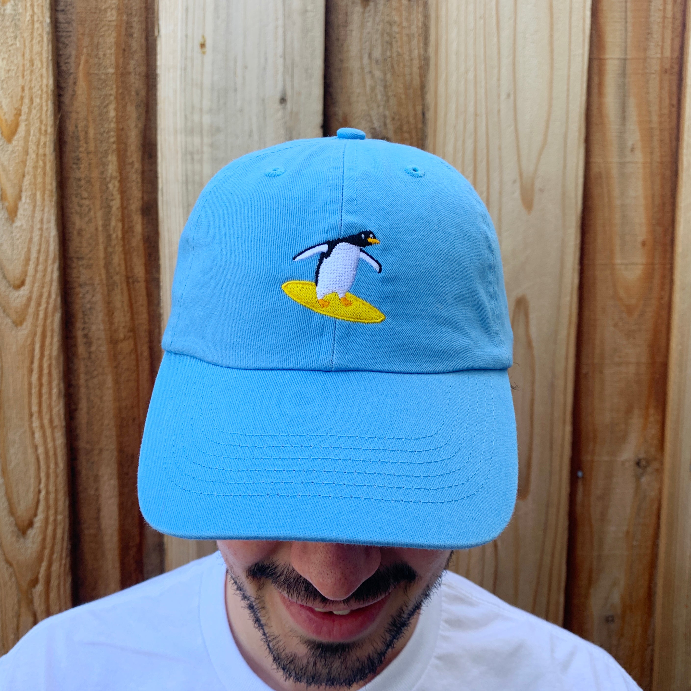 Close up of sky blue baseball cap with a surfing penguin embroidered on the front.