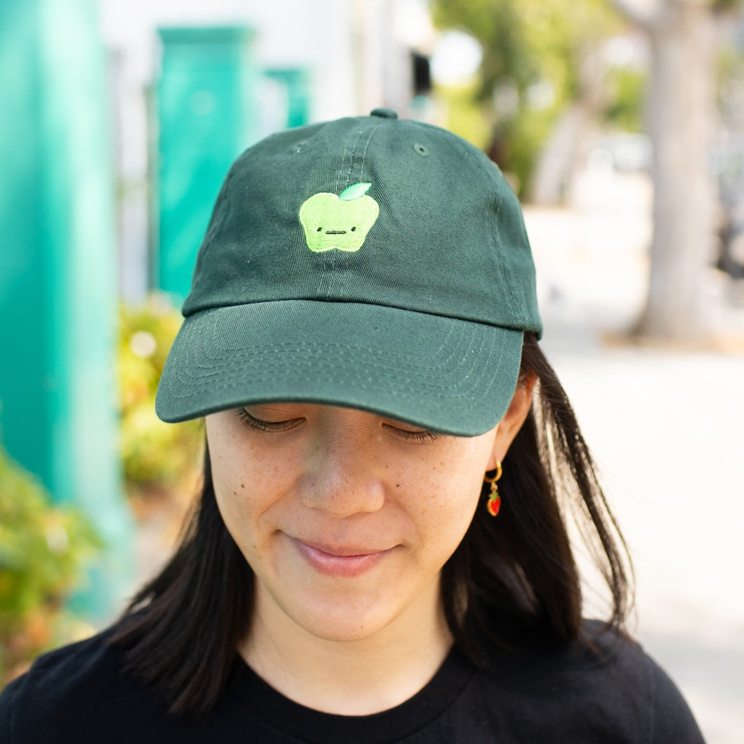 Person smiling and looking down, wearing green baseball cap with light green apple  with a straight face embroidered on front