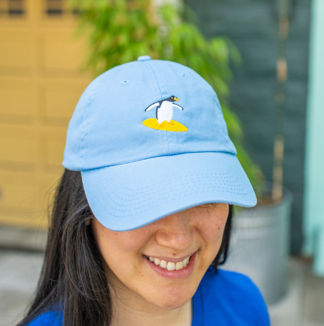 Person smiling and wearing sky blue baseball cap with a surfing penguin embroidered on the front.