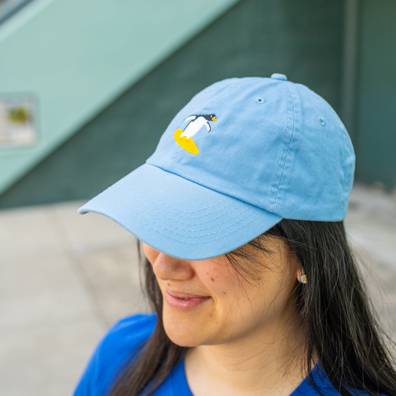 Person wearing sky blue baseball cap with a surfing penguin embroidered on the front.