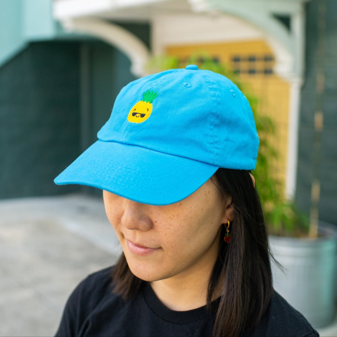 Side view of person wearing bright blue baseball cap with pineapple wearing sunglasses embroidered on the front.