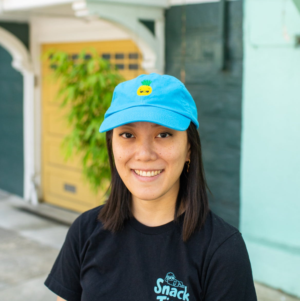 Person smiling and wearing bright blue baseball cap with pineapple wearing sunglasses embroidered on the front.