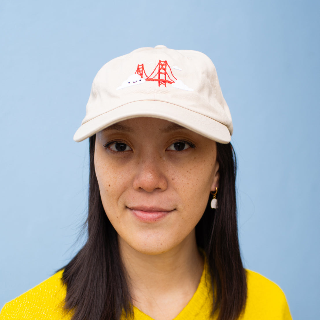 Person wearing beige baseball cap with Golden Gate Bridge and smiling clouds embroidery.