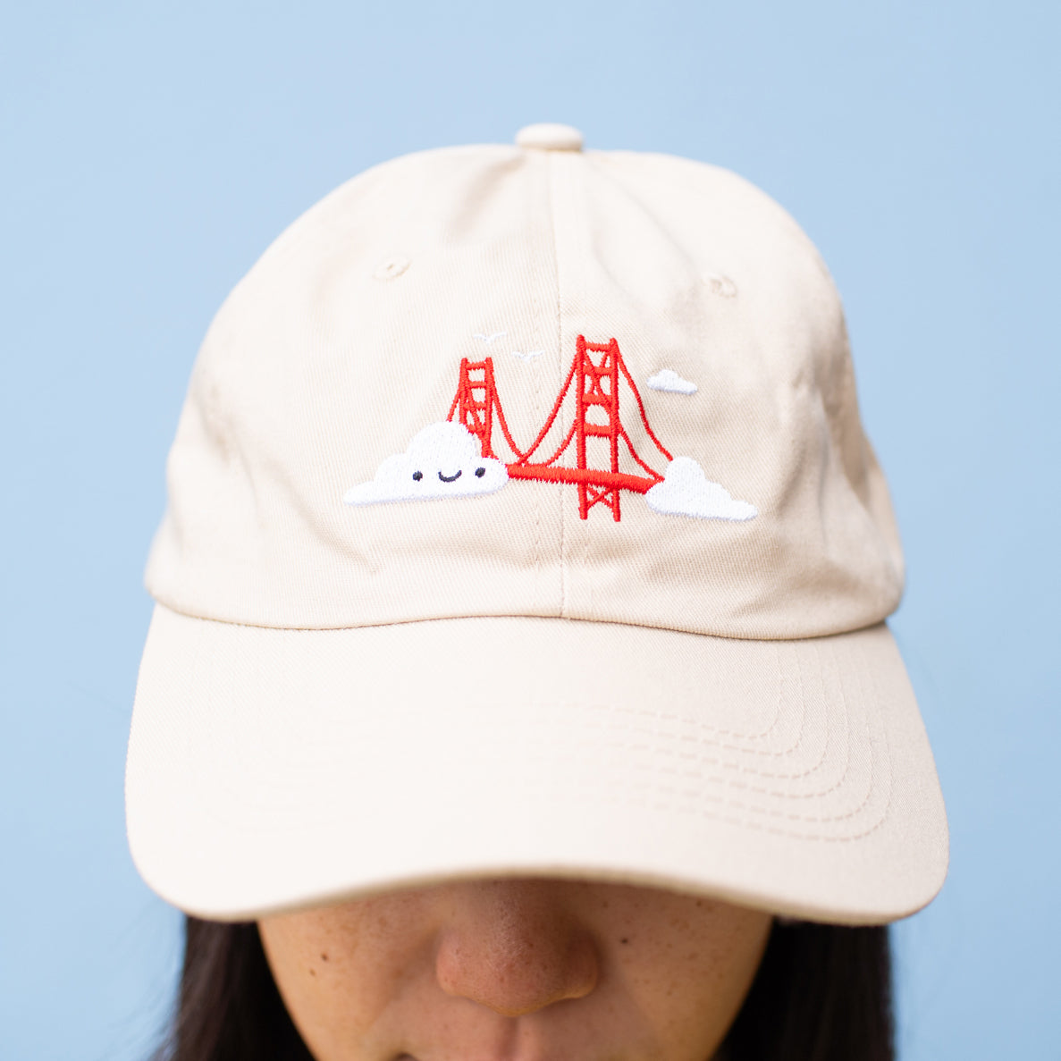 Close-up of person wearing beige baseball cap with Golden Gate Bridge and smiling clouds embroidery.