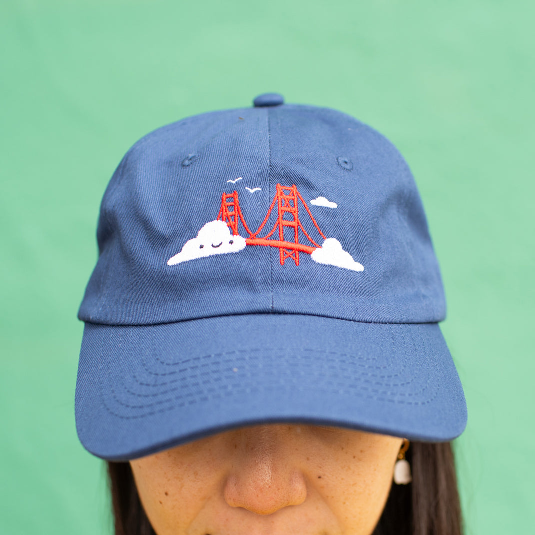 Close-up of person wearing navy blue baseball cap with Golden Gate Bridge and smiling clouds embroidery.