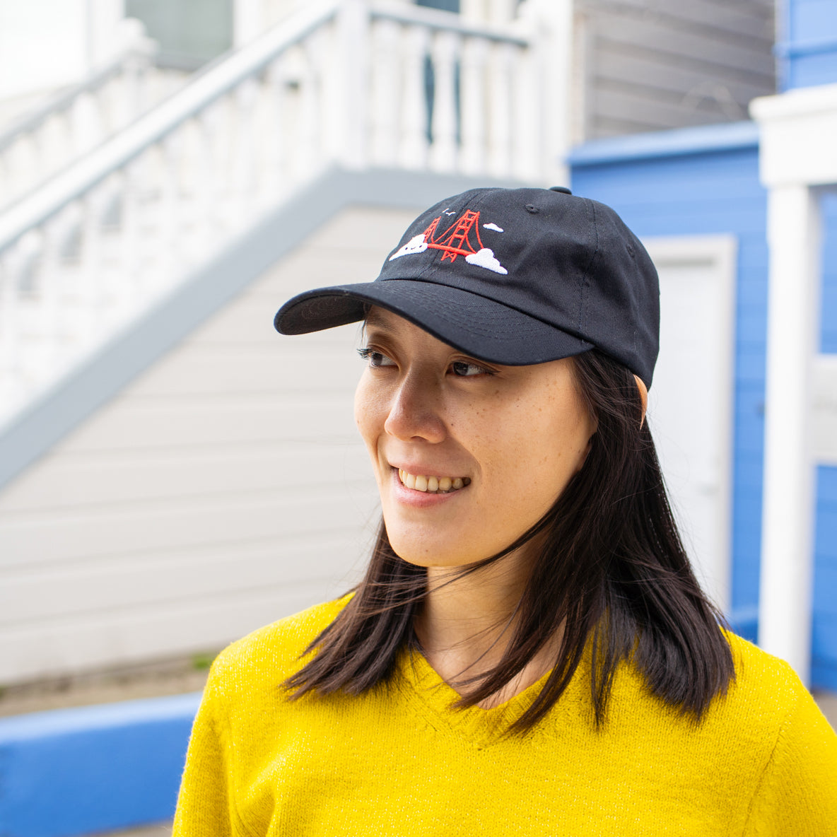 Person smiling and looking into the distance, wearing black baseball cap with Golden Gate Bridge and smiling clouds embroidery.