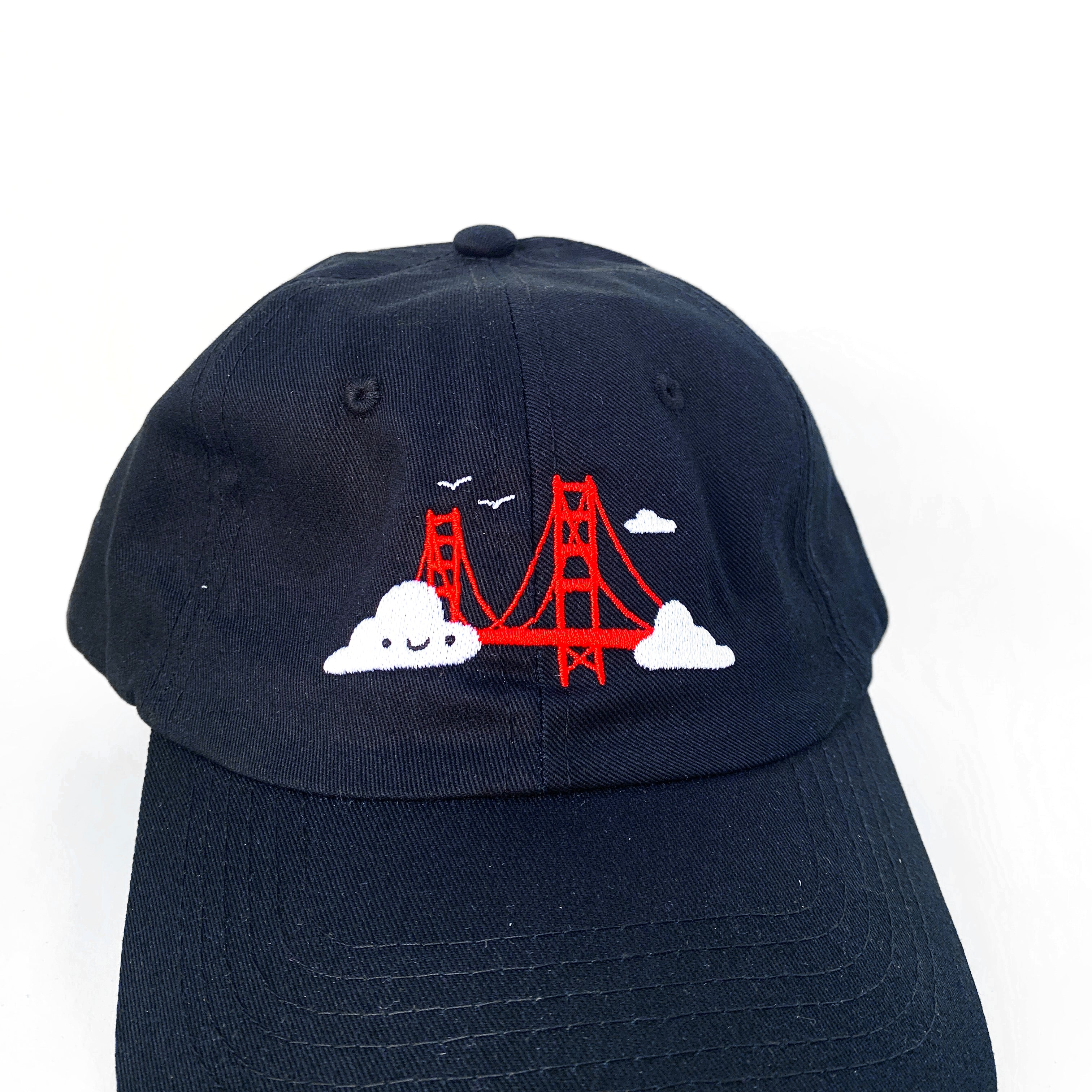 Close up of black baseball cap with Golden Gate Bridge and smiling clouds embroidery.