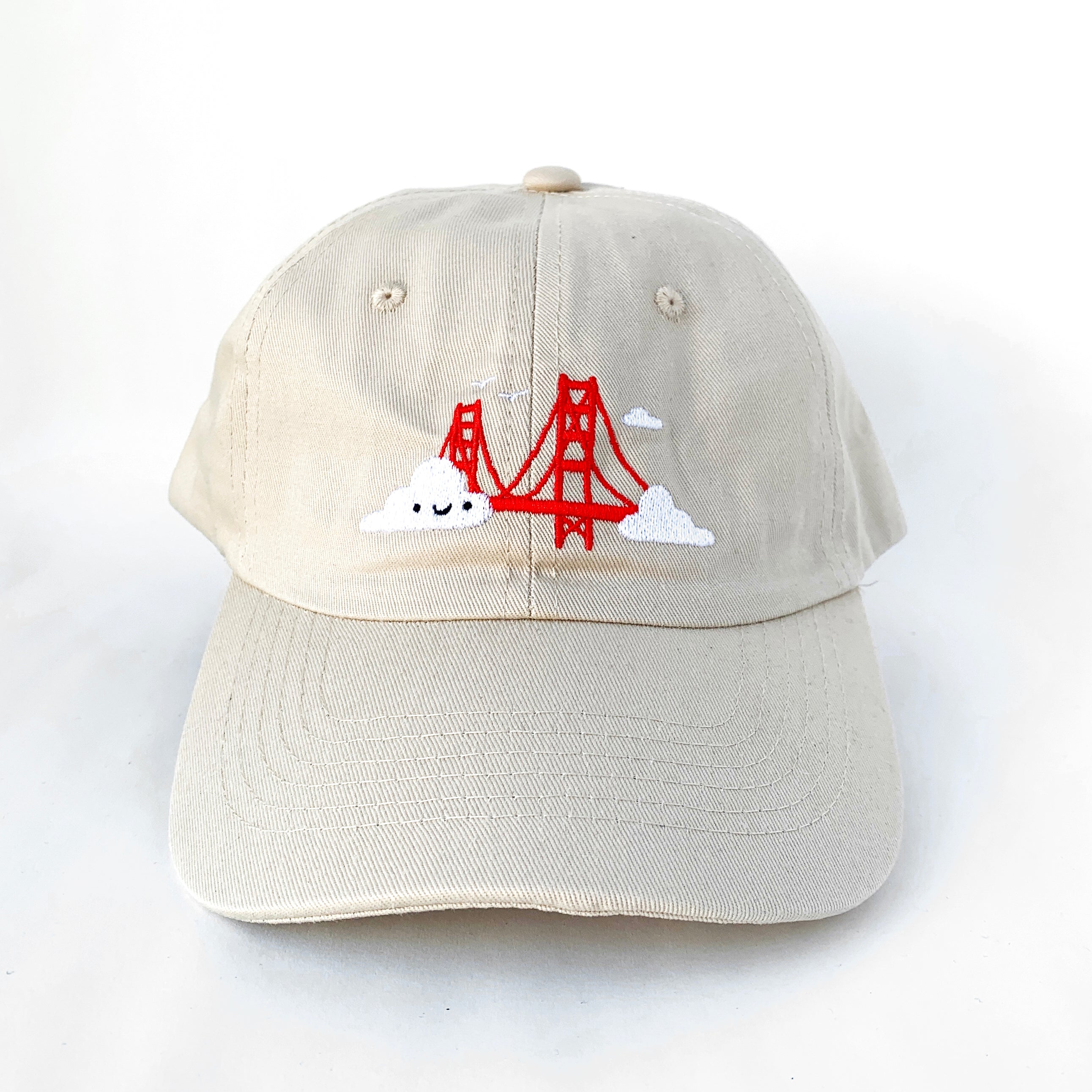 Light beige baseball cap with Golden Gate Bridge and smiling clouds embroidery.