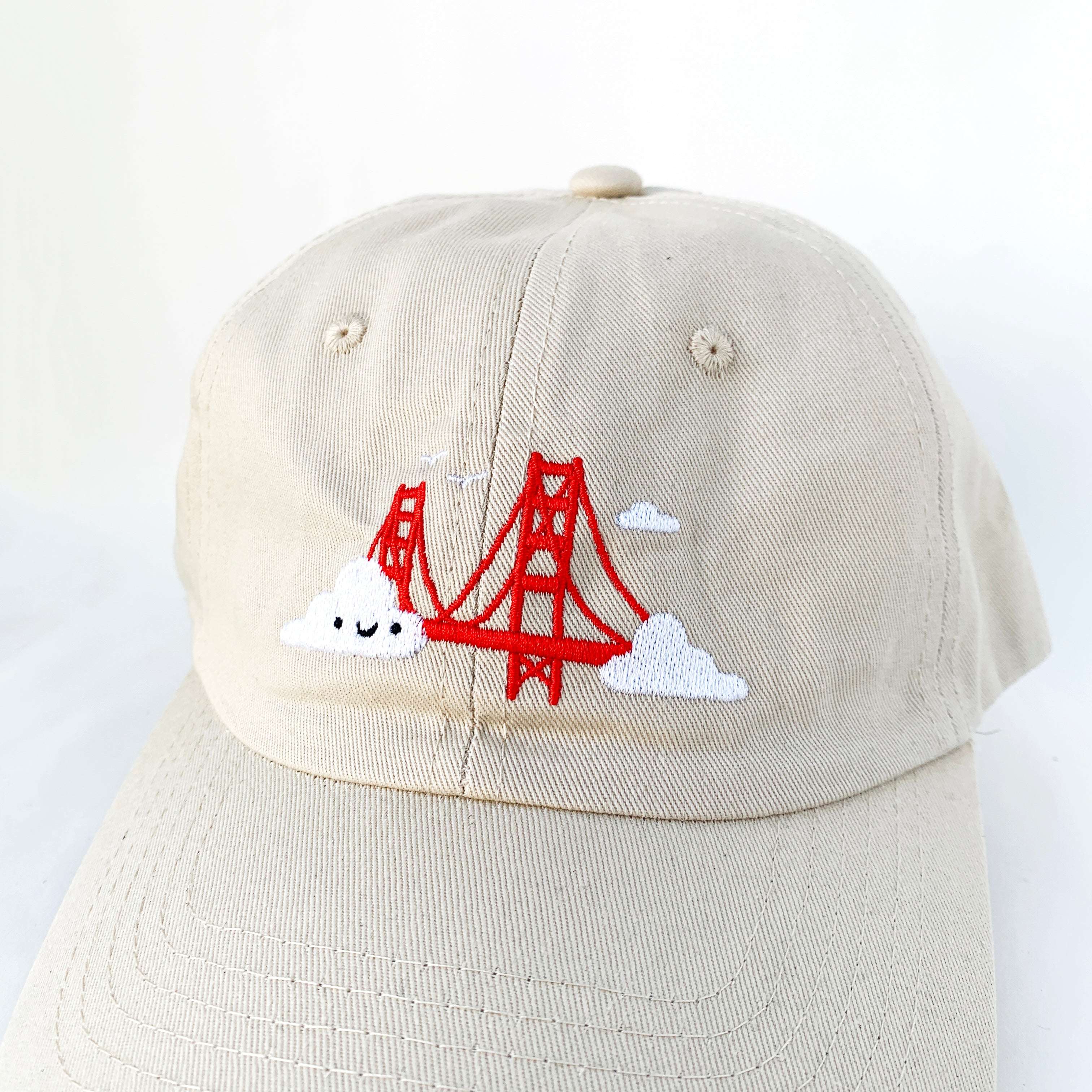 Close up of light beige baseball cap with Golden Gate Bridge and smiling clouds embroidery.