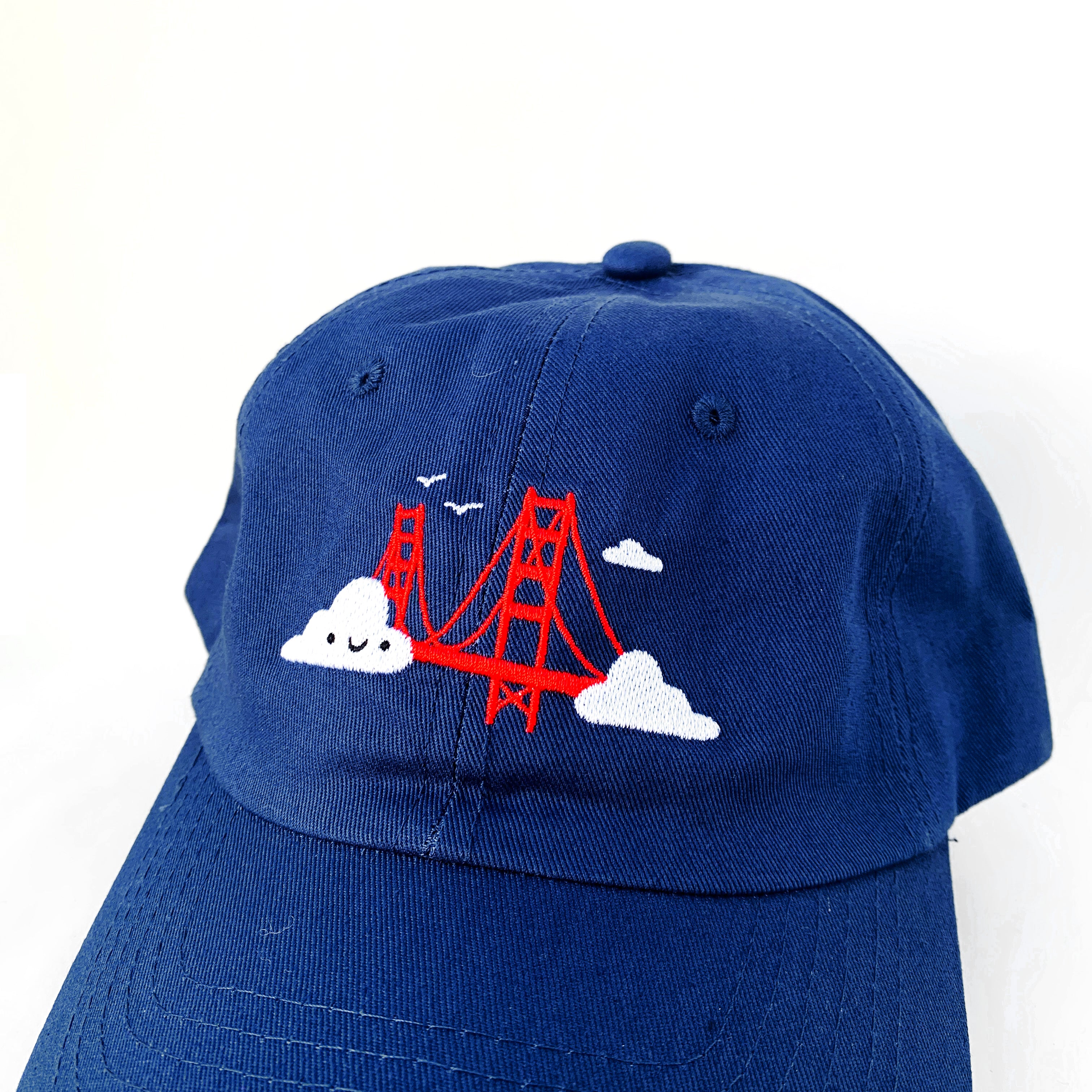 Close up of navy blue baseball cap with Golden Gate Bridge and smiling clouds embroidery.